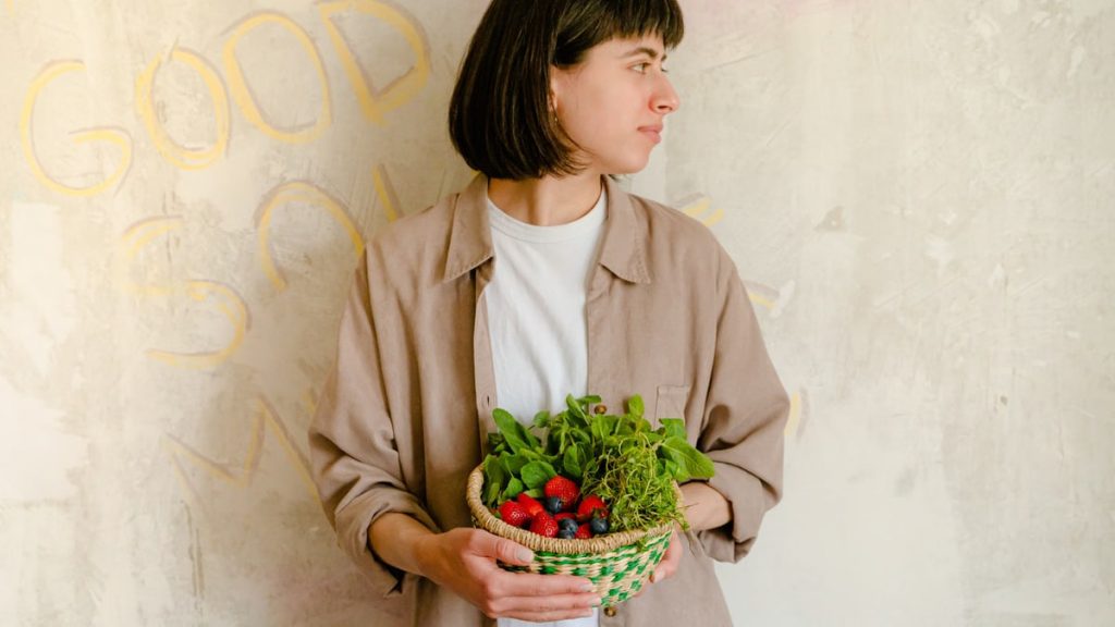Woman standing with a basket of fresh greens and berries against a rustic wall background.